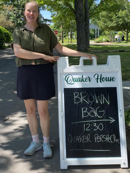 Alicia McBride standing by a sandwich board reading 'Brown Bag | 12:30 | Quaker Perspective'